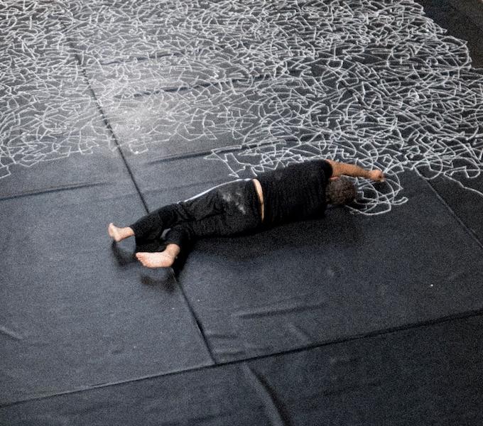 Paolo lays on a black floor drawing white lines across the surface